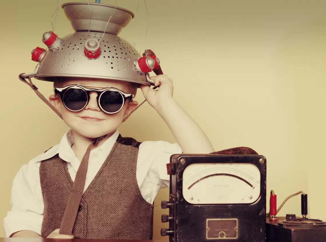 A young boy wearing goggles with a metal colander on his head sitting next to a machine 