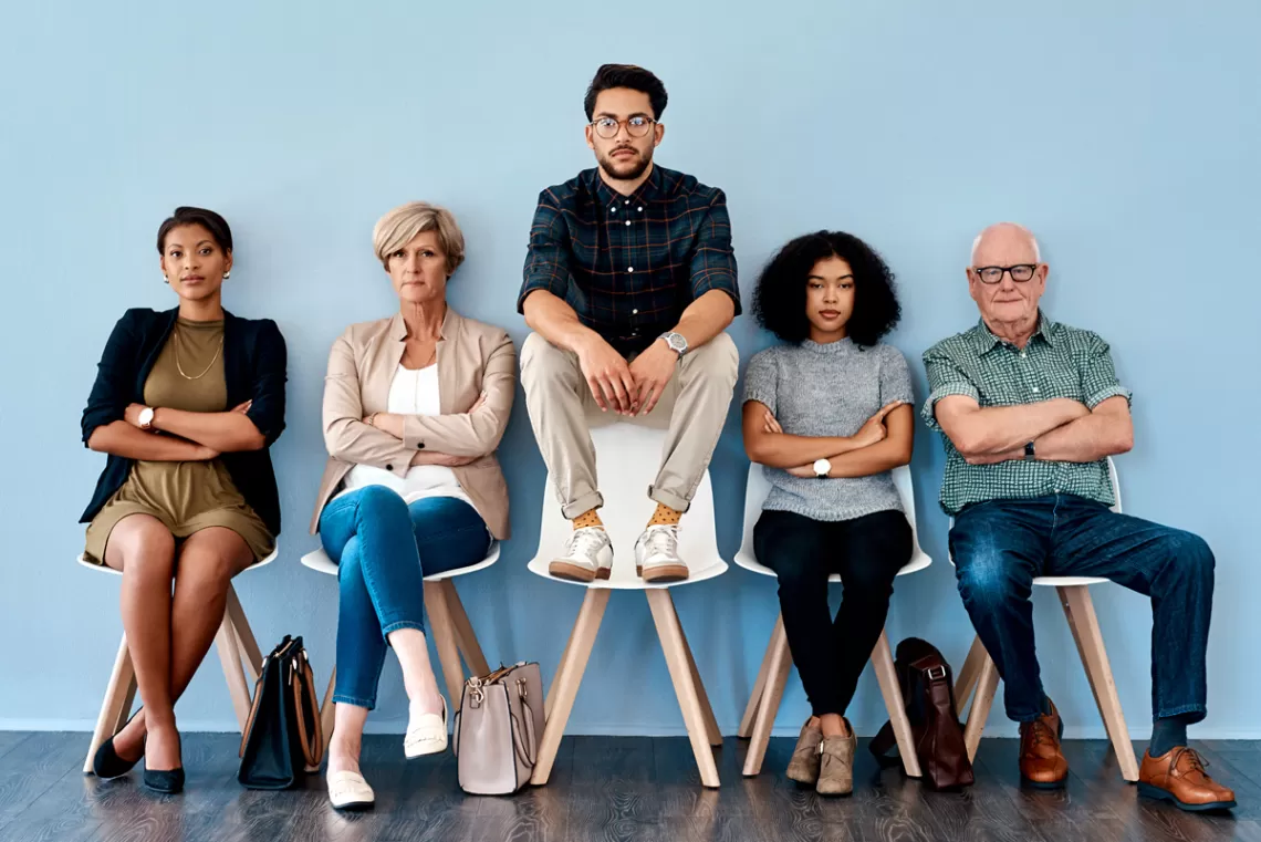 five people sitting in a row of chairs, the third person is sitting on the back of the chair instead of the seat