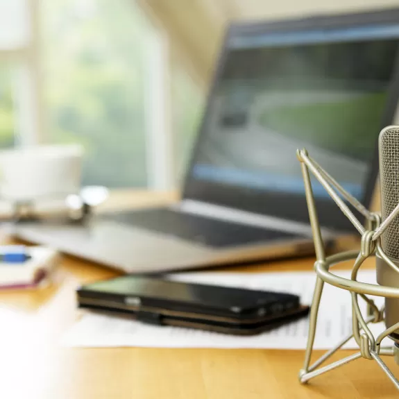 A desk set up with a microphone in the foreground and a laptop, phone, paper and pencil, glasses, and a mug