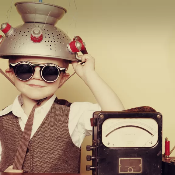 A young boy wearing goggles with a metal colander on his head sitting next to a machine 